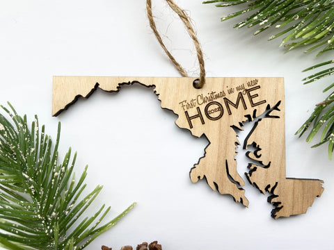 A wood ornament in the shape of the state of Maryland on a white background, surrounded by green foliage. The ornament is laser-cut with precise detail and hangs on a jute string.